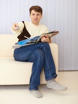 The young man sits on a sofa with the newspaper and remote control