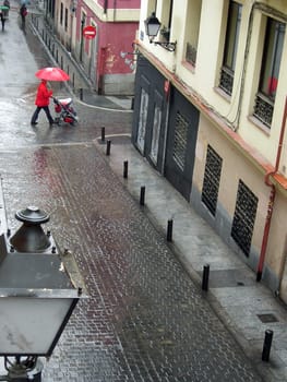 A man dress in red walking with his baby on a street of Madrid in spite of the rain.