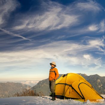 Man stand with yellow tent on snow hill against dramatic sky.