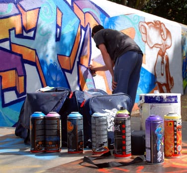 A photograph of an unidentifiable graffiti artist spray painting a public wall, with paint and cans in the foreground