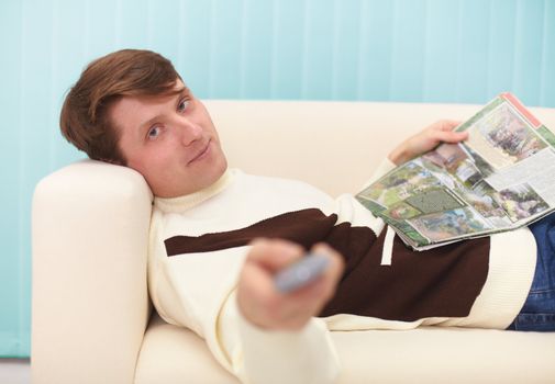 A smiling man, lying on the couch with a magazine and the TV remote control