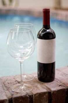 A red wine bottle and two empty glasses by the pool. Plenty of copy space on the blank wine label.