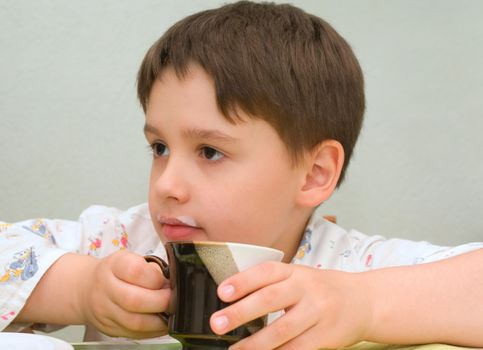 Little boy is sitting at the table with a cup and spoon. There is milk trace on his lips