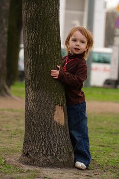 Little beauty girl playing hide-and-seek at the park. She hideing round the big old tree with split off part of bark.