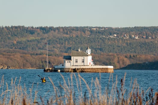 The white lighthouse in the Oslo fojrd with autumn-coloured Ekeberg in the background, lit by the setting sun.
