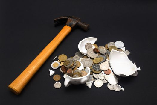 A broken piggy bank isolated on a dark background with loads of coins from around the world and a hammer.