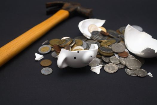 A broken piggy bank isolated on a dark background with loads of coins from around the world and a hammer.