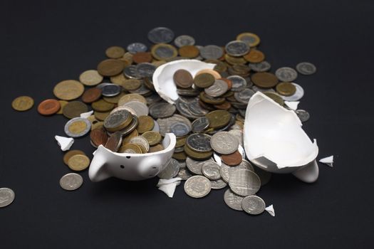 A broken piggy bank isolated on a dark background with loads of coins from around the world.