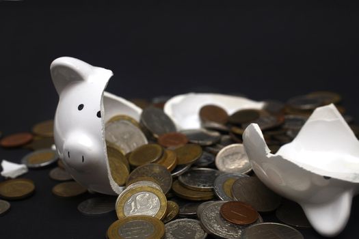 A broken piggy bank isolated on a dark background with loads of coins from around the world.