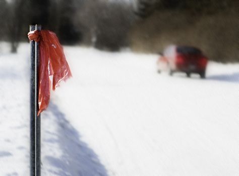 A  car having problem on a snowy, windy day.  A red cloth is tied to a pole on the left as a warning to other drivers.