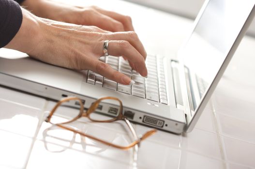 Female hands typing on the keyboard of the laptop with eyeglasses nearby.