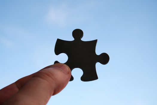 Black jigsaw piece in human fingers on a blue sky background