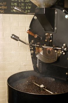 The freshly roasted beans from a large coffee roaster, just as the beans are stirred in the cooling cylinder. Smoke rises while the dark roasted beans are stirred.
