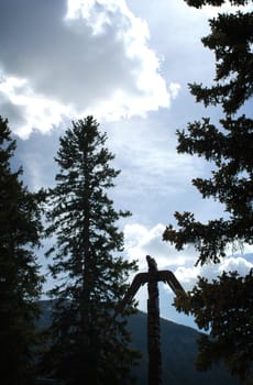 Silhouette image of a totem pole eagle between pine trees against a blue sky with clouds.