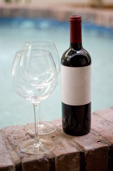 A red wine bottle and two empty glasses by the pool.  Plenty of copy space on the blank wine label.