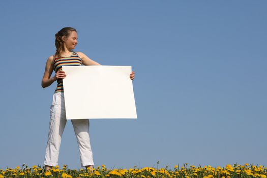 Nice girl in a flowering field, holding blank white cardboard with copy space.