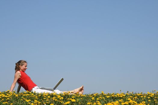 Casual young woman relaxing with laptop outdoors, in a flowering dandelion field.