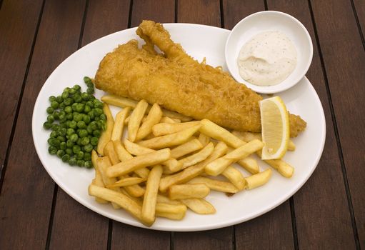 Fish and chips served with peas and tartar sauce on white plate on wooden pub table.