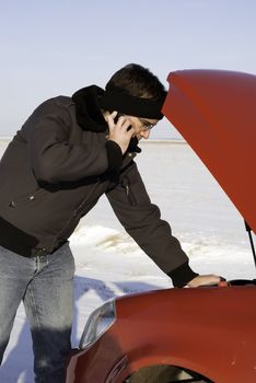 A young man is checking under the car hood while talking on a cell phone.