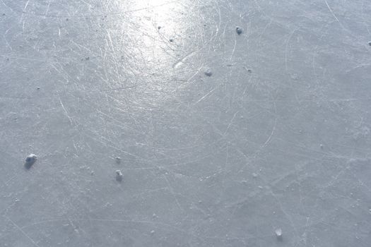 Surface of an outdoor ice rink replete with skate marks reflects the sun.