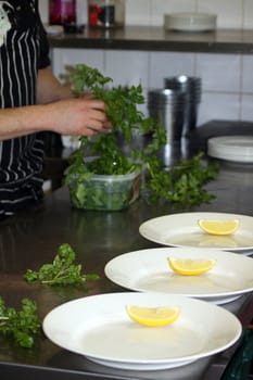 Preparation of food in a restaurant kitchen. Three white oval plates being dressed in readiness for service with wedges of lemon and fresh watercress.