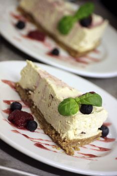 A slice of freshly made chees cake set on a round white plate with fresh summer fruits topped with a sprig of fresh basil. set at an angle.