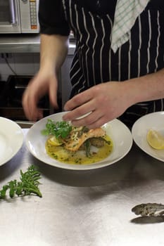 A chef in a restaurant kitchen dressing a plate of freshly cooked salmon over a bed of new potatoes with fresh watercress.