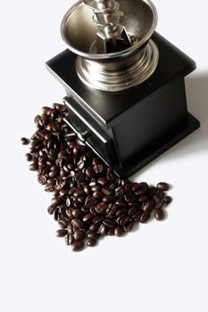 A portrait format image of a black wooden hand coffee grinder set amongst a pile of coffee beans, set on a white isolated background.