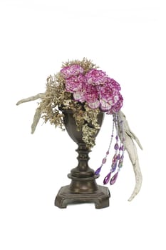 A floral arrangement of dried materials and pink carnation flowers set on a white isolated background. Pink and purple costume jewellery cascading from a bronzed vessel with pink carnations set with dried mosses and wood.