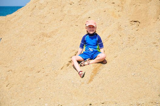 happy little blond boy wearing surfing suit is sitting in the huge pile of sand on the beach, good weather, summer is here! 