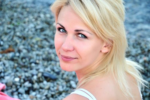 portrait of  beautiful young blond woman with stunning blue eyes, she is sitting on the 	 pebble near the sea.