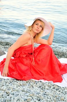 beautiful young woman wearing white hat, strand of pearls and red dress is sitting on the beach near the sea