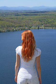 Red-haired girl looking over blue lake from the mountain.