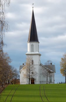 A wooden church staying on a hillock, overa a grass field, with an alleyway to one side. The church is situated in Fet, Norway.
