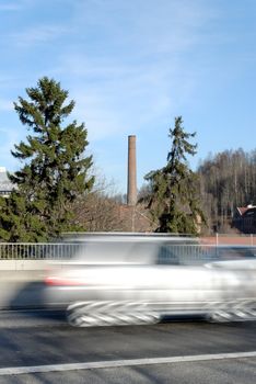 A silver car zooming by on a highway bridge, set against a old factory chimney. Shot at Nydalen, Oslo, Norway
