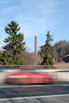 A red car zooming by on a highway bridge, set against a old factory chimney. Shot at Nydalen, Oslo, Norway
