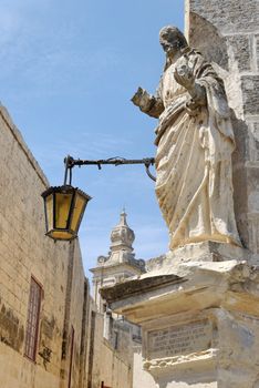 Eroded standstone statue of St.Peter on a corner of a street in Mdina, Malta. The sign's inscription dates back to 1860. Carmelite church is seen in the background
