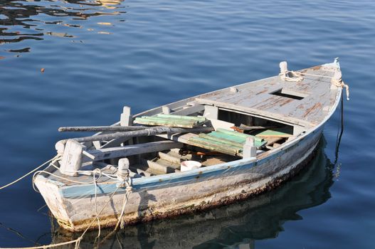 Old wooden fisher boat tied to buoys (not visible) in a Croatian harbor. Inside the boat you can see a wooden mast and paddles plus fishing equipment. The water has a great blue tone and some reflection on tiny waves.