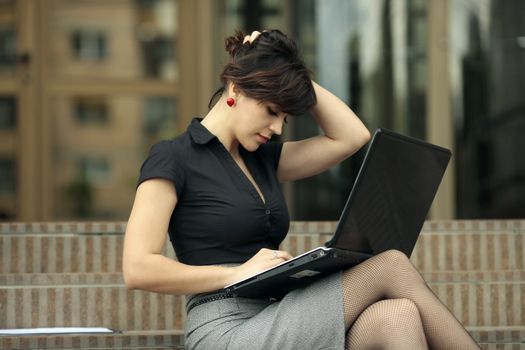 busy young attractive lady playing with her hair and looking at a laptop on the stairs in front of an office building