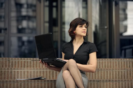young attractive lady with a laptop on the stairs in front of an office building looking away