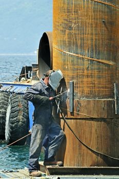 Worker using soldering iron on a rusty pylon in a harbour