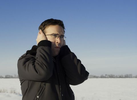 A young man standing outside in the winter, holding his ears to keep them warm