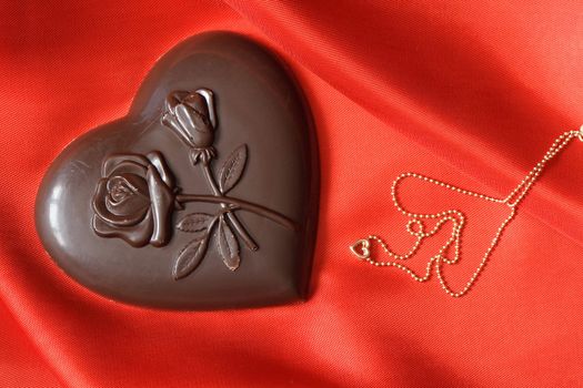 Closeup of chocolate heart and gold necklace lying on red silk textile with copy space