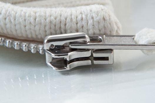 Close up and low level angle of a metal zip slider attached to cream coloured knitwear arranged on a white reflective surface.