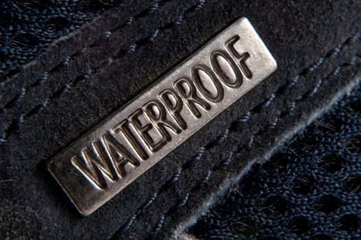 Close up of a small metal badge engraved with the word 'waterproof' and attached to dark blue suede material with dark stitching.