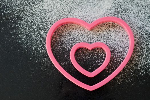 Two heart shaped cookie cutters on a granite worktop, with a dusting of icing sugar