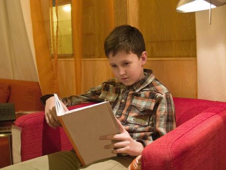 The boy sitting in a red armchair and reading the book