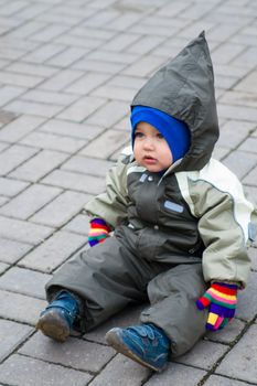 1 year old boy in green snowsuit with multicolored gloves sitting  on paving stone