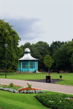 A park's green space with trees and grass, with a bandstand and a dog running with a ball.