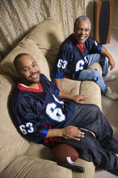 Portrait of an African-American father and son sitting on couch smiling and wearing football jerseys.